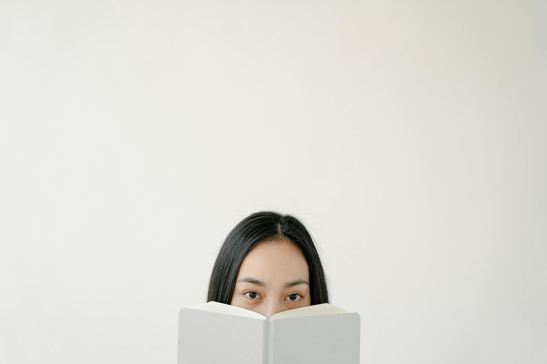 Nieśmiała kobieta chowająca twarz za książką na tle pustej ściany