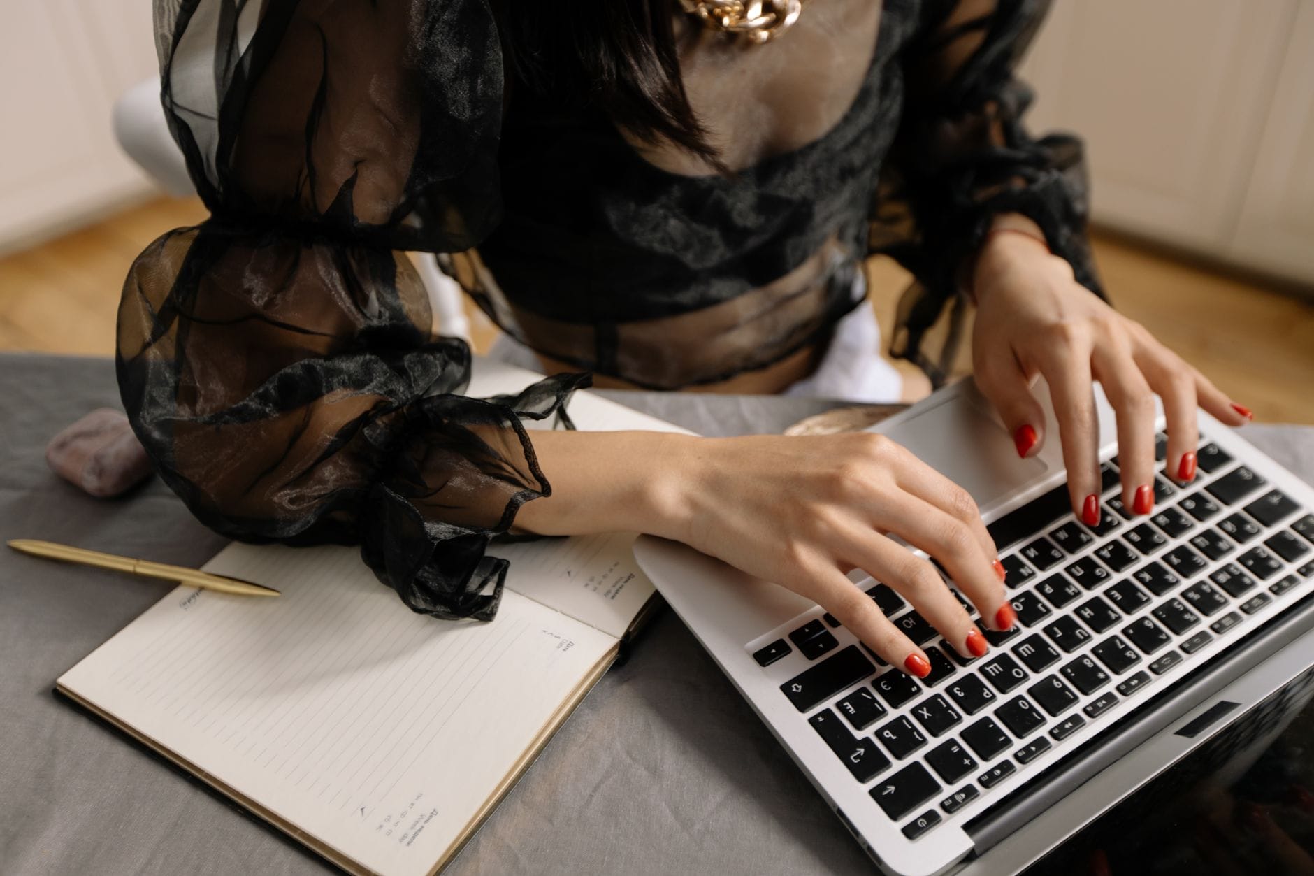 Kobieta z czerwonymi paznokciami pisze na laptopie obok notesu